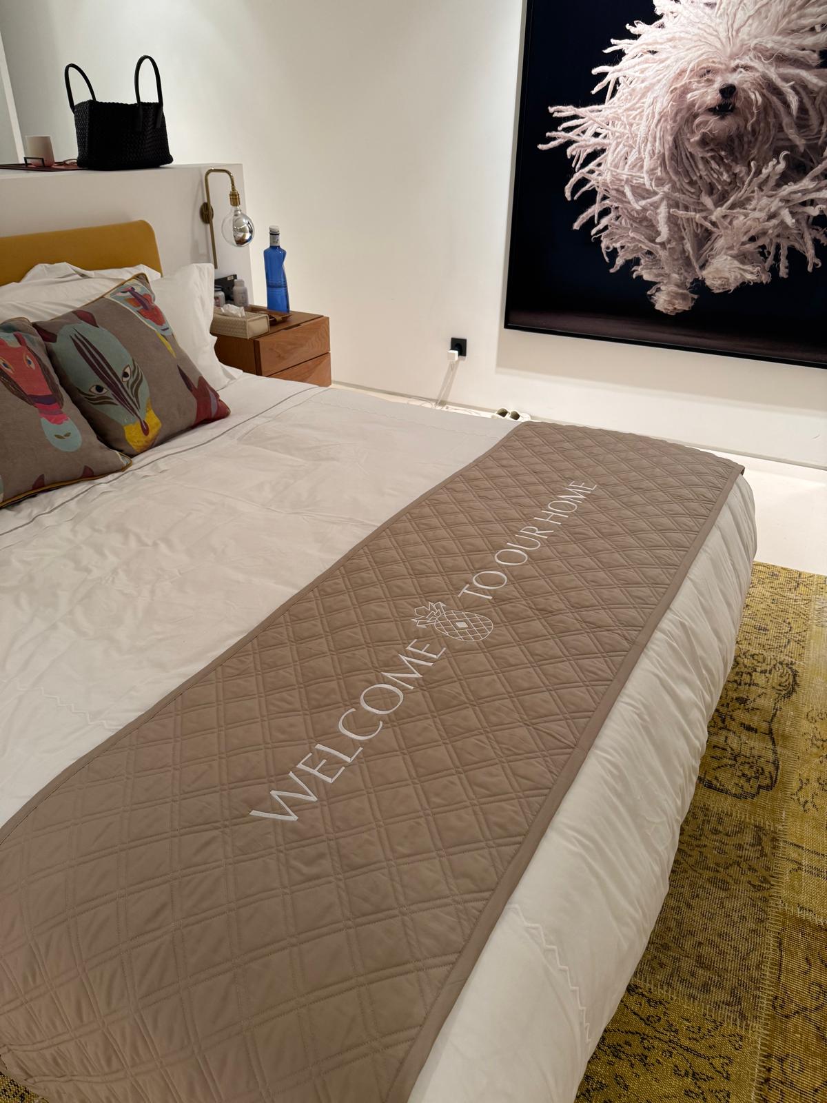 Neatly made bed with a quilted runner that reads “Welcome to our home"