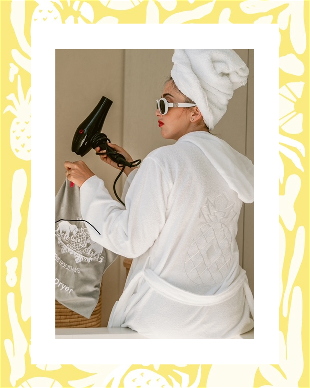 Woman in a white robe and towel turban holds a hairdryer and bag, wearing sunglasses with red lipstick. Background has a playful pineapple border.