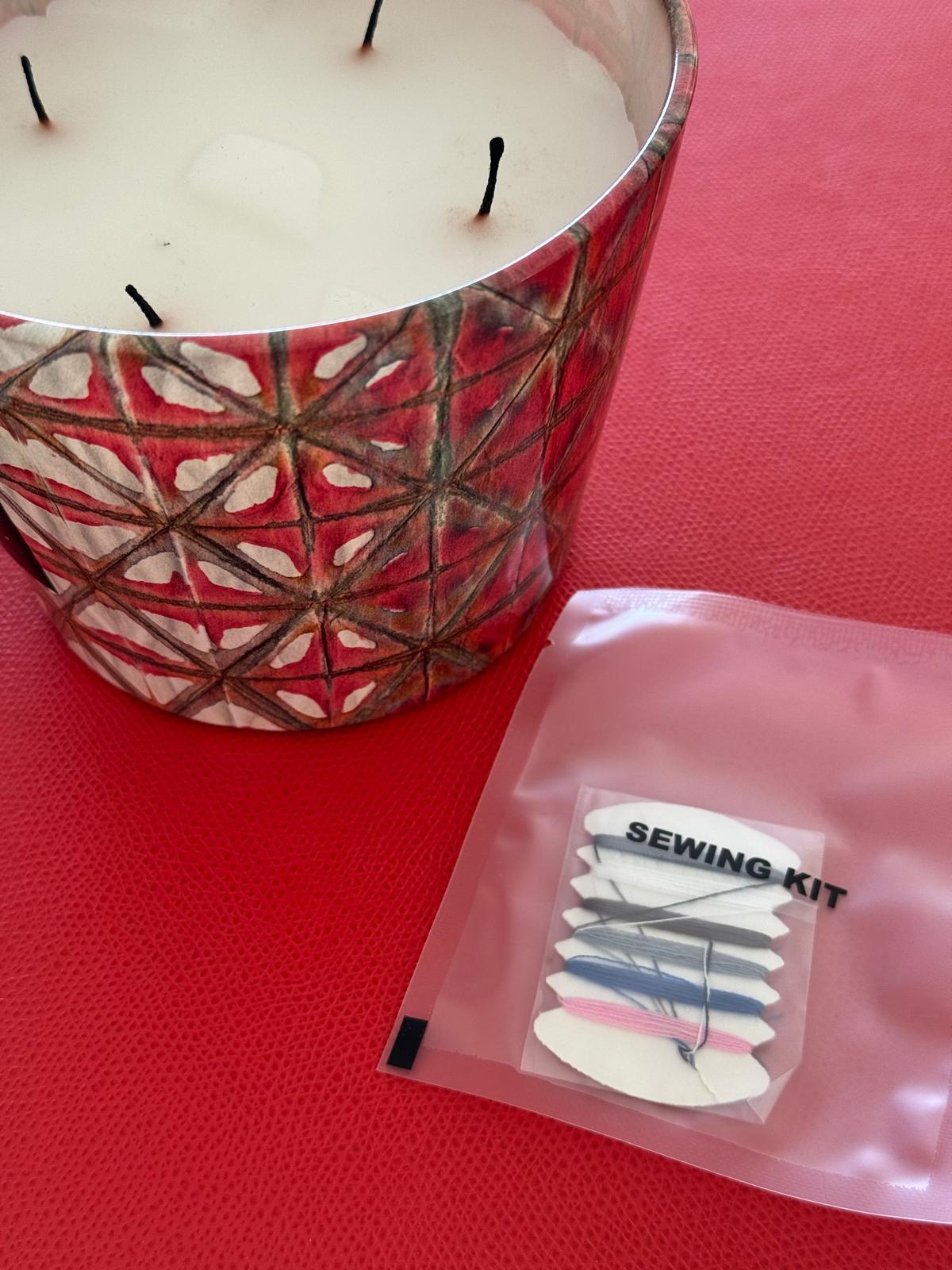 Candle with geometric pattern next to a small sewing kit on a red surface.