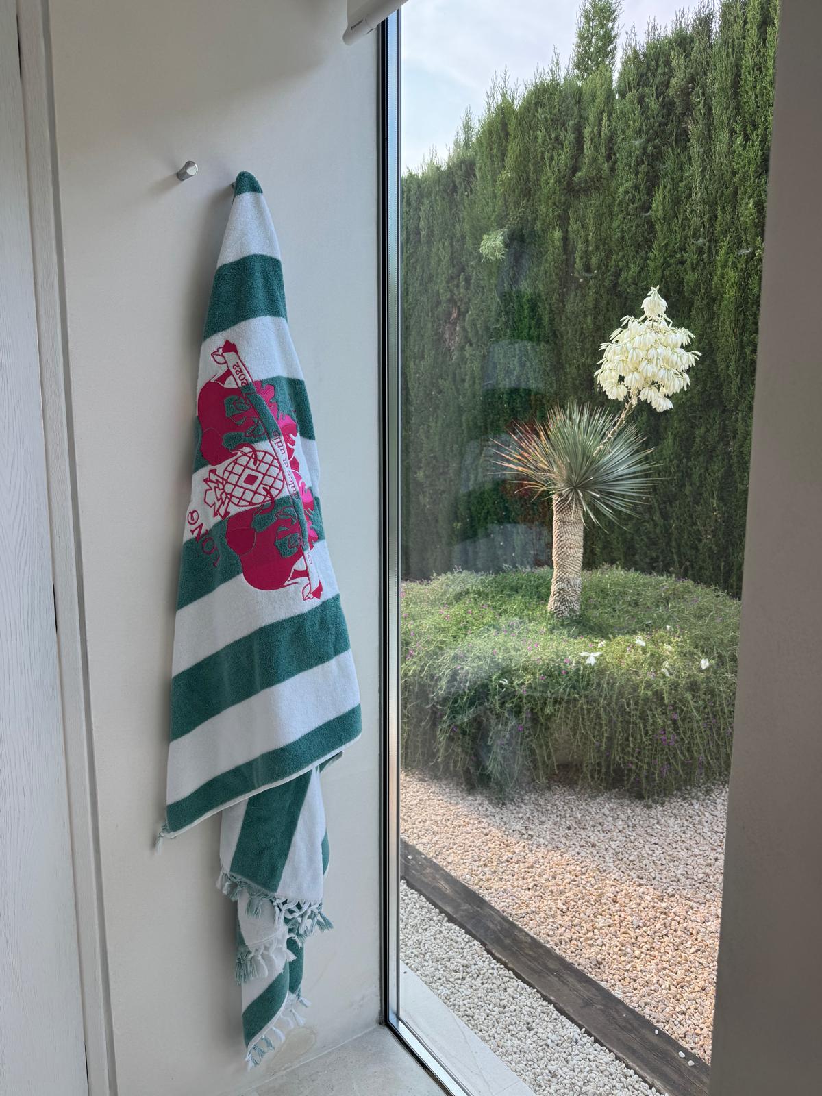 A striped towel hangs on a wall hook beside a window with a view of a garden. Outside, a yucca plant blooms near a hedge and gravel path, creating a serene setting.