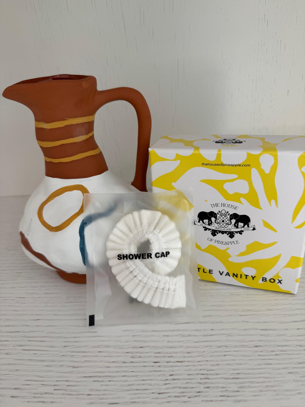 Shower cap in packaging placed in front of a ceramic jug and a yellow-and-white vanity box.